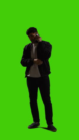 Vertical-Video-Full-Length-Shot-Of-Young-Man-Talking-On-Mobile-Phone-Standing-Against-Green-Screen-With-Low-Key-Lighting-2
